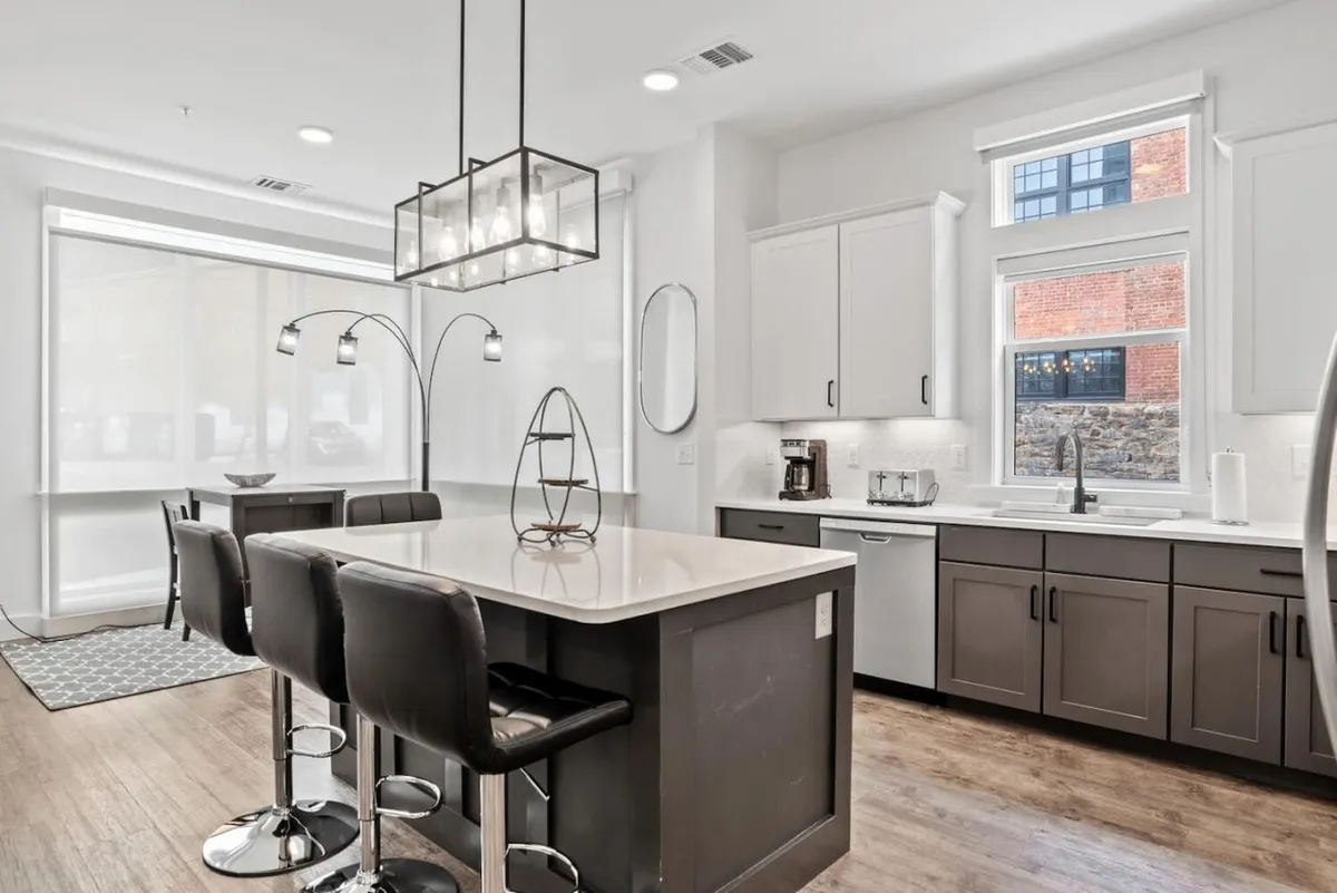 A bright kitchen scene with a spacious island, modern light fixtures, and bar stools for casual dining. Large windows bring in natural light, and the open layout connects seamlessly to adjacent living spaces, making it ideal for entertaining or relaxed meals.