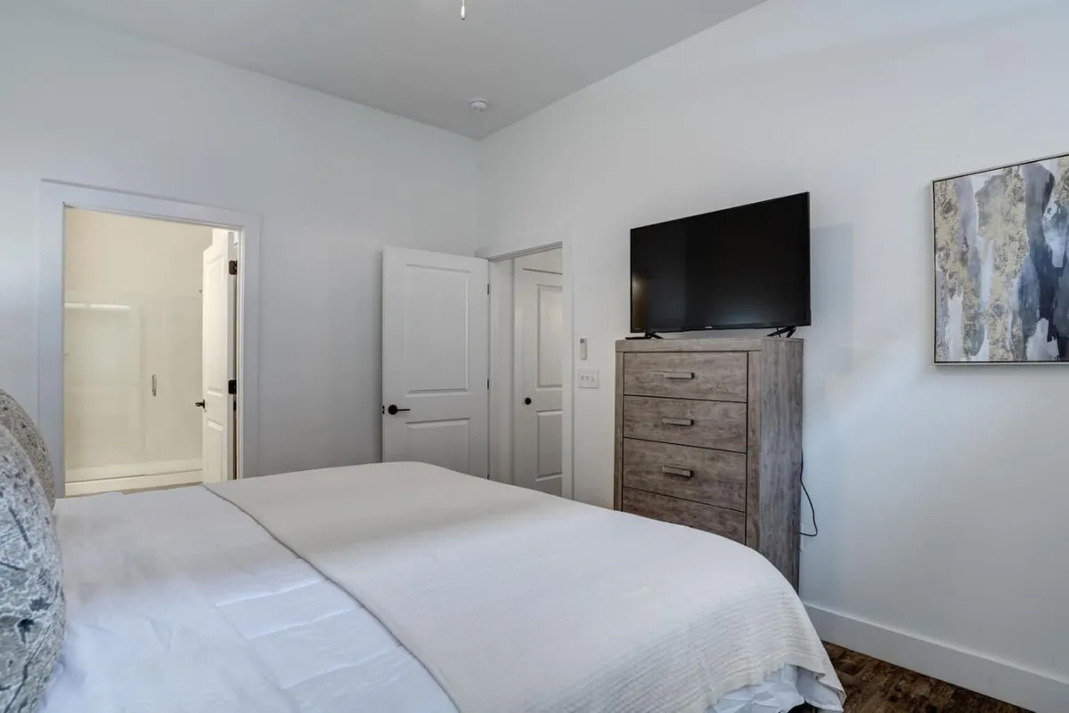 A stylish bedroom with cozy bedding, a dresser, wall-mounted TV, and unique artwork. The room exudes a serene mood, perfect for winding down with a movie after a busy day or enjoying a quiet morning in bed.