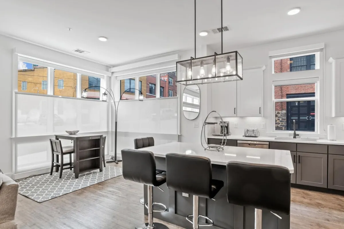 A spacious, light-filled open-plan area featuring a kitchen island, dining table, and large windows. The contemporary light fixture and the presence of both prep and dining spaces suggest a setting welcoming for group activities, meals, and socializing. The abundant natural light emphasizes the bright, airy atmosphere.