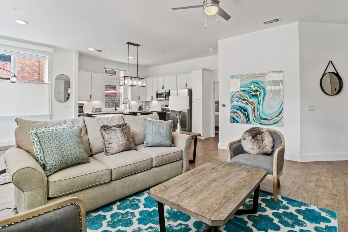 A comfortable living room scene with a sofa, coffee table, and visible kitchen area in the background. Modern artwork and thoughtful lighting complete the look, showcasing a stylish yet relaxed environment ideal for gatherings and shared experiences.