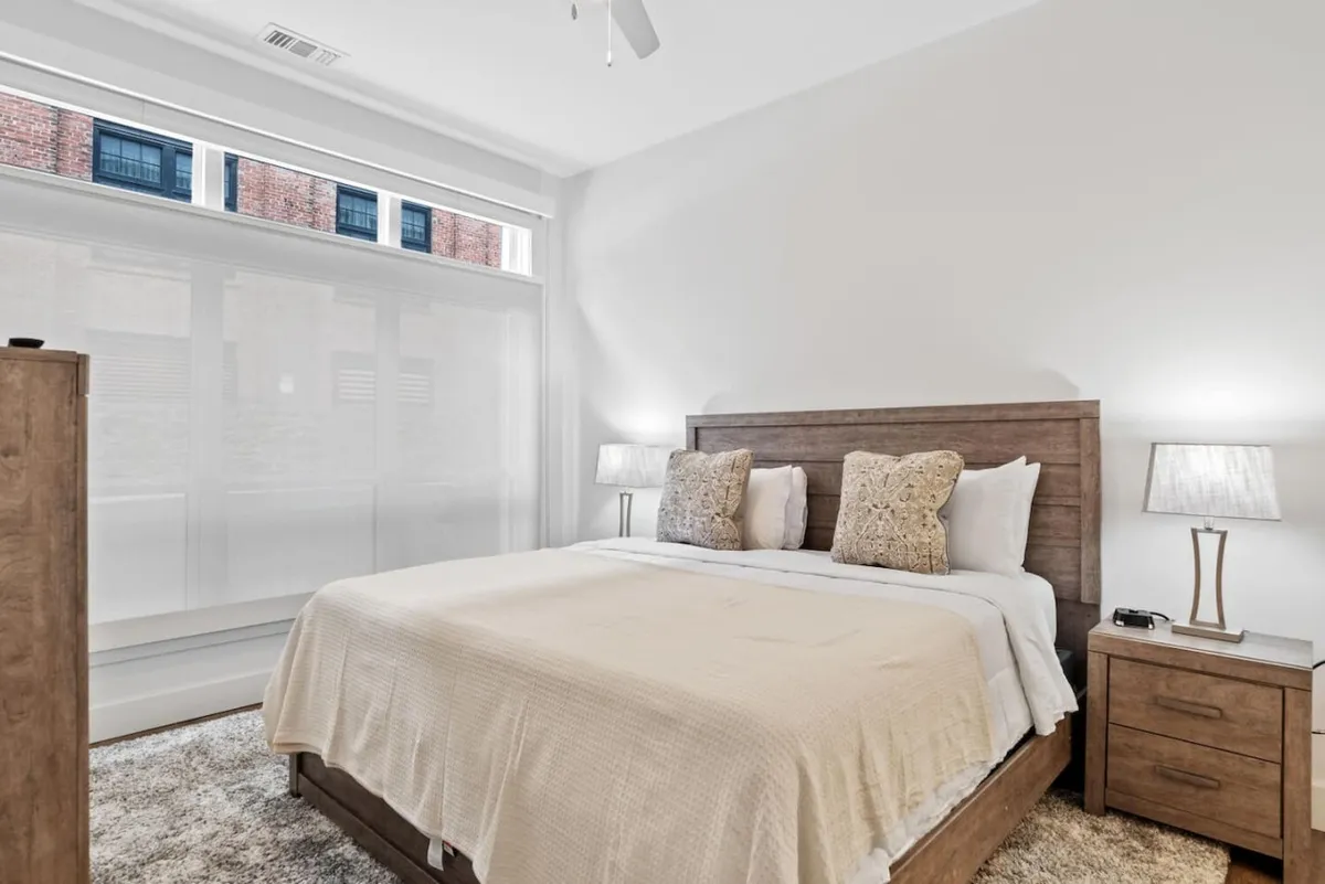 Featuring a spacious king bed adorned with decorative pillows and flanked by stylish nightstands and lamps, this room is bathed in soft, inviting light that creates a tranquil, sophisticated atmosphere ideal for relaxation.