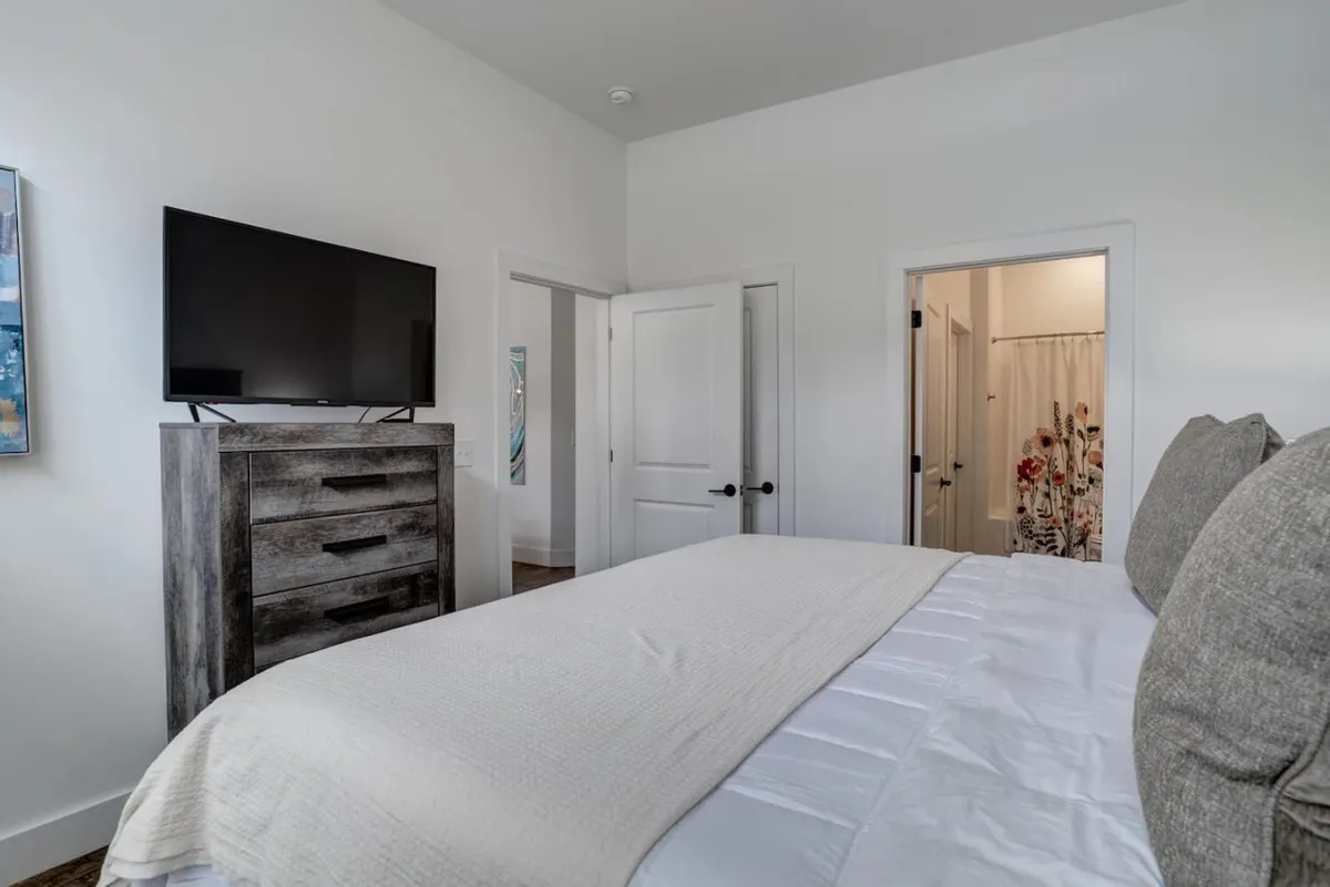 A cozy bedroom scene with a bed featuring white bedding, a television, a wooden dresser, and doorways leading to other rooms. The atmosphere is comfortable and inviting, suggesting a relaxing retreat after experiencing the city.