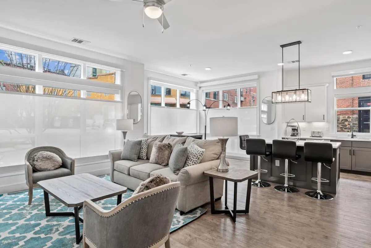 The image showcases a sunlit open-concept living area featuring a cozy sofa, a modern coffee table, a spacious kitchen island for dining, and expansive windows that bathe the space in natural light. The layout is perfect for relaxing with loved ones, enjoying a cup of coffee by the window, or sharing meals together after a day exploring Asheville.