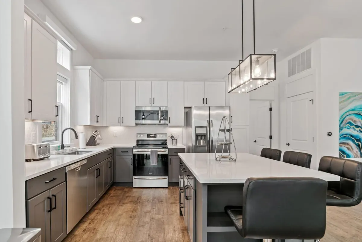 A modern kitchen featuring a central island surrounded by stainless steel appliances and accented by contemporary light fixtures. Colorful wall art adds a playful touch, while the spacious layout offers ample room for cooking and gathering. Bar stools at the island suggest a space for casual dining or conversation.